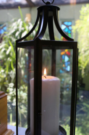 Image is a white candle burning; behind it, the greenery from the atrium can be seen through the sanctuary windows.