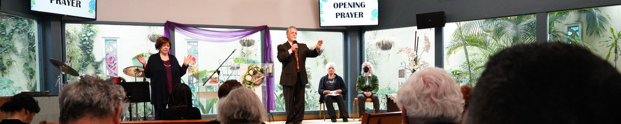 Image shows our sanctuary during a Sunday service. Rev Greg is giving an opening prayer while our lead prayer chaplain, Michelle, raises her hands while holding space for the prayer. A lush, green atrium shows behind a wall of windows at the back of the stage. Two large screens show a slide that says "Opening Prayer."