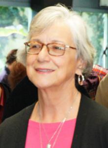 The image is a woman smiling. She is wearing glasses, a pink shirt, and a black jacket.