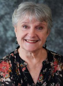 Image is a smiling woman with short, gray hair and a floral shirt.