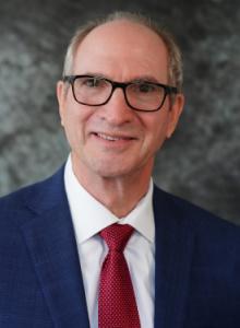 Image is a smiling man with short hair and glasses, wearing a blue suit, white shirt, and red tie.