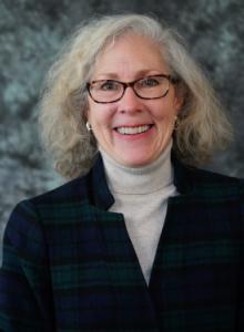 Image is a smiling woman with medium-length gray hair and glasses. She is wearing a blue and green plaid jacket over a tan turtleneck.
