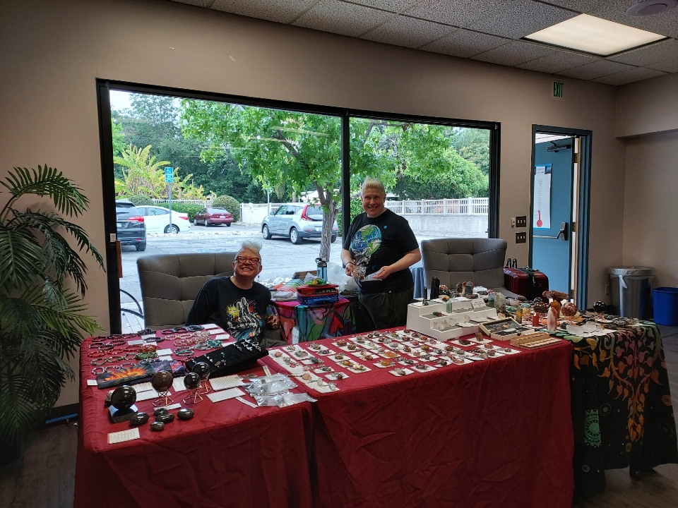 Image shows a table with lots of different crystals displayed. Steph and Sarah Darshan from Lighthouse Crystals are smiling.