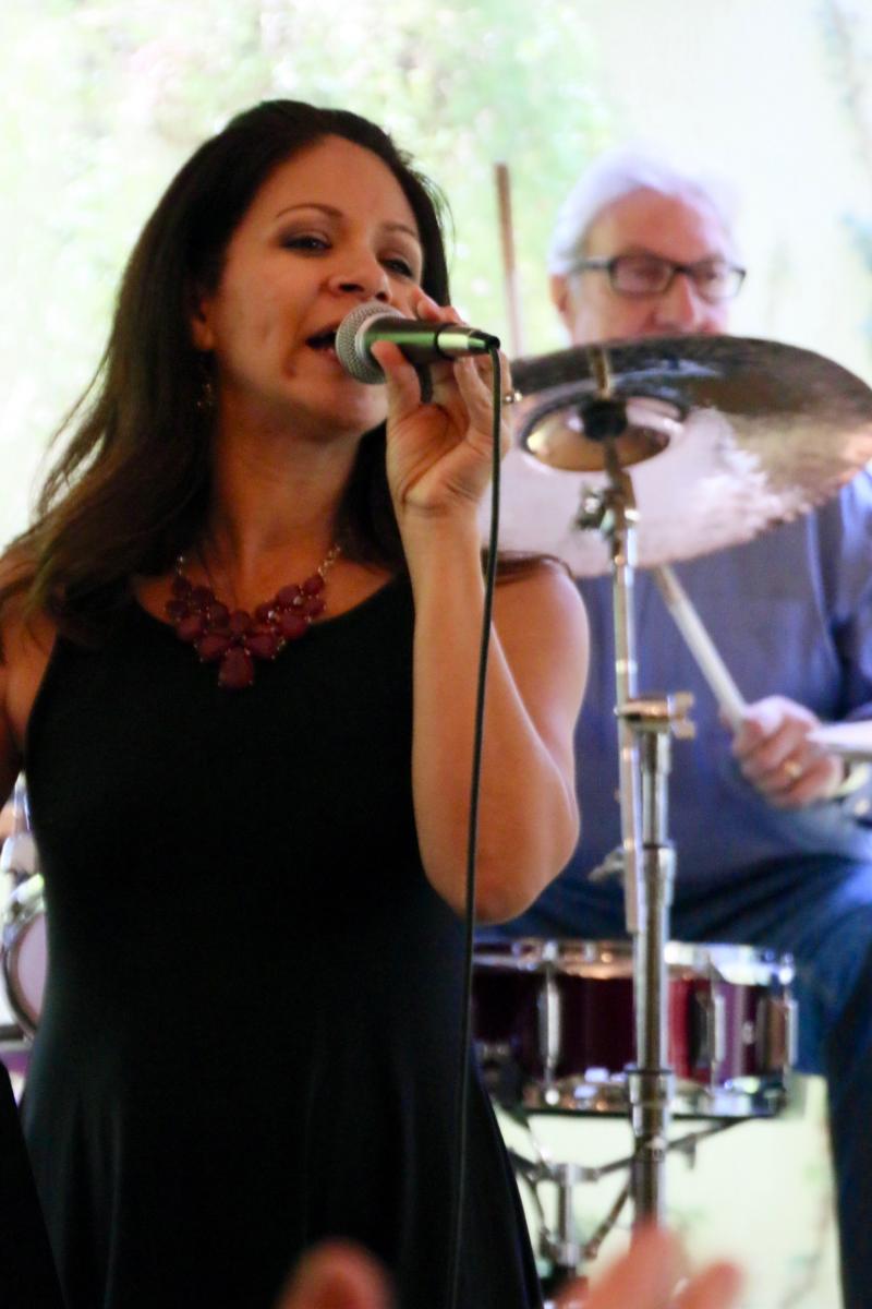 Image is Felice Hernandez-Schaeffer singing into a microphone. Behind her, Gary Clason plays the drums.