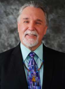 Image is a headshot of Rev. Greg Dorst wearing a suit and tie, and prayer beads