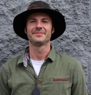 Image is a headshot of Johnny Sage, smiling, wearing a green shirt, brown hat, and a sun pendant.