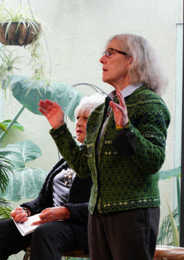 Image is two women at the front of the sanctuary. One is standing with her hands out in prayer; the other is seated behind her.