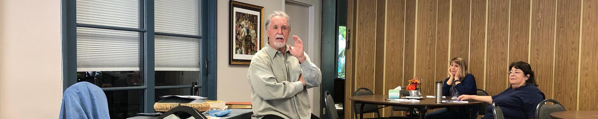Rev Greg stands in front of a group of people sitting at tables in fellowship hall.