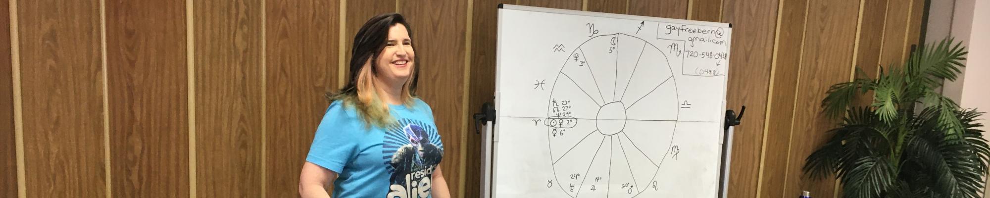 Picture shows teacher Gay Freebern standing by a whiteboard with an astrology chart drawn on it.