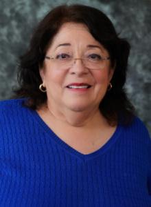 Image is a woman with medium-length brown hair and glasses, wearing a bright blue sweater.