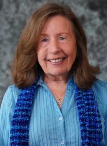 Image is a smiling woman with medium-length brown hair, wearing a light blue shirt and a darker blue scarf.