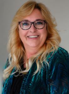 Image is a smiling woman with long, blond hair and glasses. She is wearing a turquoise jacket over a black shirt.