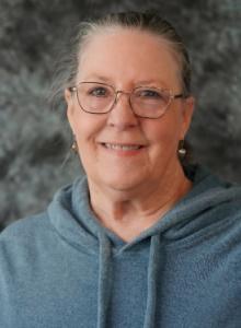 Image is a smiling woman with gray hair pulled back, wearing glasses and slate blue hoodie sweater.