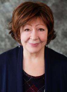 Image is a smiling woman with brown hair, wearing a blue jacket over a plaid top.