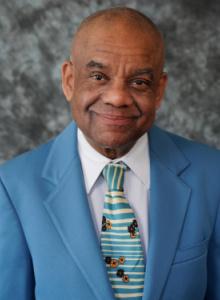 Image is a smiling man with a shaved head, wearing a light blue suit jacket over a white shirt and colorful tie.