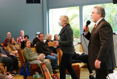 Image is Rev Greg speaking into a microphone during a Sunday service as an usher prepares to hand out a donation basket to the people in the pews.